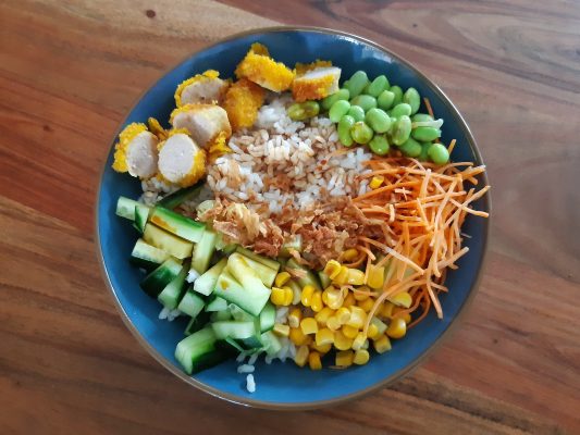 pokebowl met krokante kip, sojabonen, wortels, komkommer en sushi rijst in een kom van bovenaf gezien.