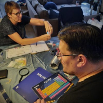 Op de voorgrond zie je een man puzzelen met een loep en politiedossier en op de achtergrond zie je een vrouw een proefje doen. Allemaal spelmaterialen uit Voorhovens Vennootschap van Codekrakers