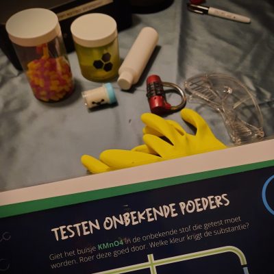 tekst met 'testen onbekende poeders' en op de achtergrond handschoenen en diverse potjes met poeders en pilletjes. Dit zijn spelmaterialen uit Voorhovens Vennootschap van Codekrakers
