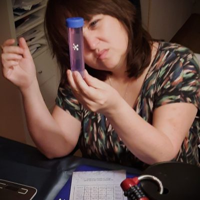 Cynthia met een buisje in haar hand terwijl ze nadenkend kijkt. Ook zie je een slot, weegschaal en papier met puzzels